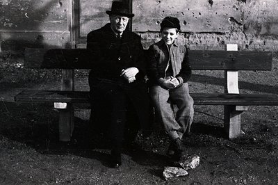 A formally dressed older man with a bowler hat sits beside a young boy on a weathered park bench. The background features a r...