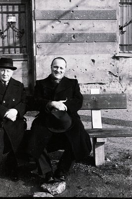 Two men seated on a weathered stone bench before a building with visible bullet holes. The man in the foreground wears a dark...
