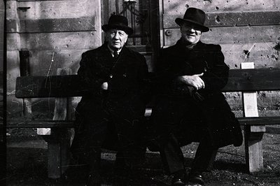 Two men in dark coats and hats sit side-by-side on a weathered wooden bench. The bench faces a large stone building, visible ...