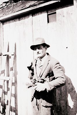 Man in a suit and fedora stands beside a weathered wooden structure, holding a bouquet of cut flowers. Appears to be a rural ...