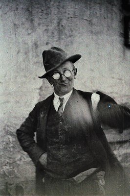 A man in a suit, hat, and spectacles poses outdoors, hand on hip. Likely 1920s or 30s, the style suggests a confident, prospe...