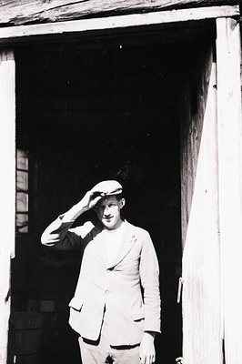 A man in a newsboy cap, blazer, and trousers stands in a doorway, tipping his hat. Appears to be a candid portrait, possibly ...
