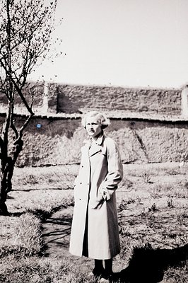 A woman in a light-colored trench coat stands in a sparse, grassy field before a weathered stone wall. Short, styled hair and...