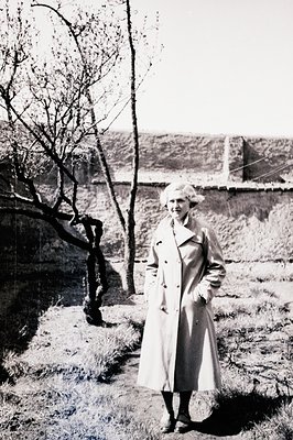 Elegant woman in a trench coat stands near a blossoming tree, posed in front of a stone wall and expansive landscape. Likely ...