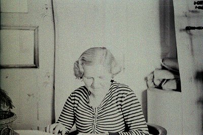 A seated woman in a striped top gazes down, appears to be writing or working on a surface. Simple interior with a framed pict...