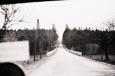 A grayscale image depicts a rural road leading toward a distant, undefined structure. The road, slightly elevated and curving...