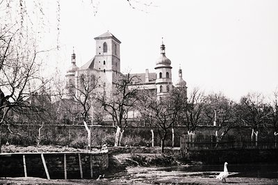 A striking black and white image depicts a grand Baroque-style church and palace complex. A low stone wall and sparse trees f...