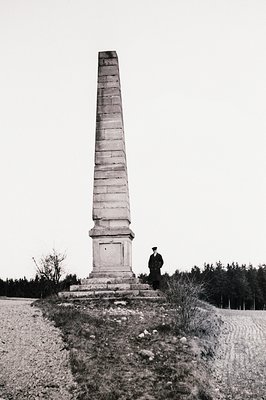 A tall, monumental obelisk stands prominently in a field, likely marking a historical event or person. A man in a suit and ca...