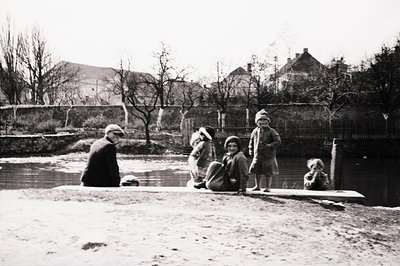 Group of children playing near a pond or stream, possibly a park or estate. Formal landscaping with stone walls and mature tr...