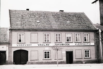A two-story building with a steeply pitched roof and a sign reading "VODODY VACLAV JAKUBICEK KLEMPIHSTVI". Features a large w...