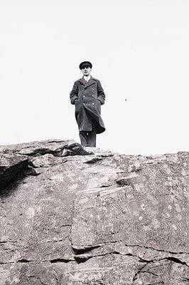 A man in a dark overcoat and cap stands prominently atop a rocky outcrop. The viewpoint is low, emphasizing the height and sc...
