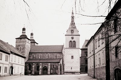 A view of a stone church and adjacent buildings within a walled courtyard. The church features a tall spire, arched windows, ...