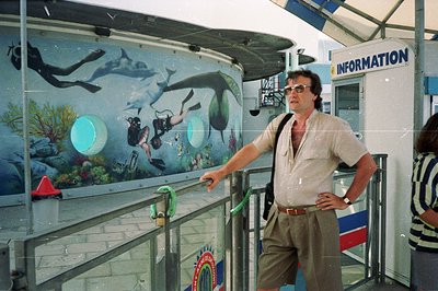 A man in 1970s leisurewear poses before a mural of divers and marine life within an enclosed structure. Signage indicates an ...