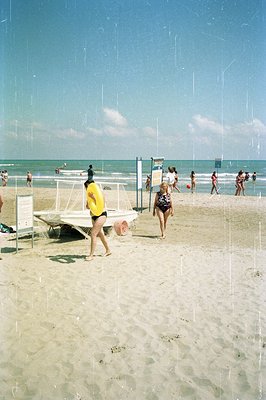 A sunny beach scene featuring swimmers and beachgoers. A lifeguard station sits prominently near the shore, with people wadin...