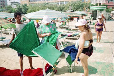 A vibrant beach scene, likely Southern Europe. A beach attendant adjusts a green sunshade while a woman with blonde hair and ...