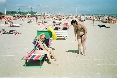 A crowded beach scene, likely in Eastern Europe. Two women are featured; one reclines on a vibrant green-and-red striped beac...