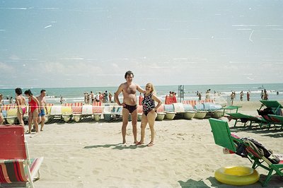 A man and woman stand on a crowded beach, likely in Bulgaria, 1970s. Classic swimwear styles and vintage beach equipment evok...