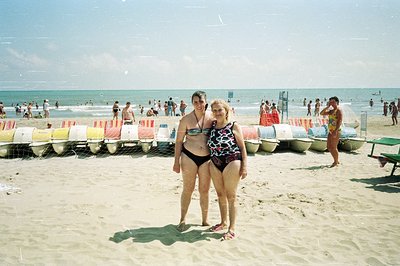Two women stand on a sandy beach, framed by a row of brightly colored pedal boats. Crowded shoreline & turquoise water sugges...