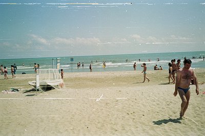 Seaside beach scene with numerous bathers enjoying a sunny day. A man walks toward the viewer in blue swim trunks; lifeguard ...
