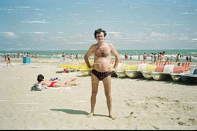 A man with dark hair, a full beard, and visible chest hair poses on a sandy beach in front of a lifeguard station. Multiple b...