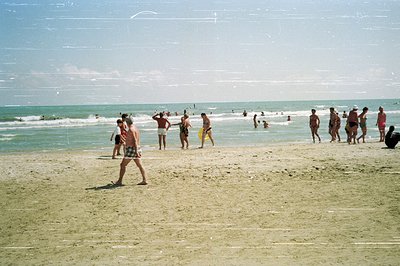 A wide shot captures a bustling beach scene, likely in Eastern Europe. Numerous people swim and sunbathe on the sandy shore a...