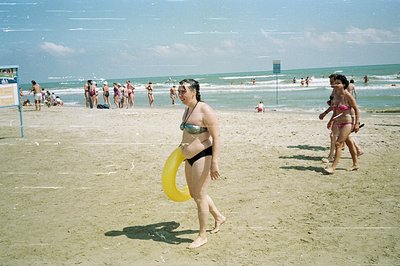 A beach scene showing two women walking along a sandy shore. One carries a yellow inflatable ring. A crowded beach and ocean ...