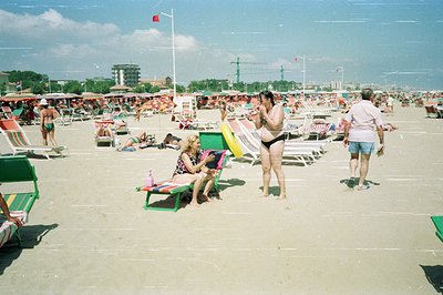 A bustling seaside scene; rows of colorful beach chairs line the sandy shore. Several people relax or walk along the beach. A...