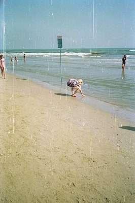 A girl in a patterned dress runs along a sandy beach toward the water’s edge. Several bathers are visible in the shallow sea ...