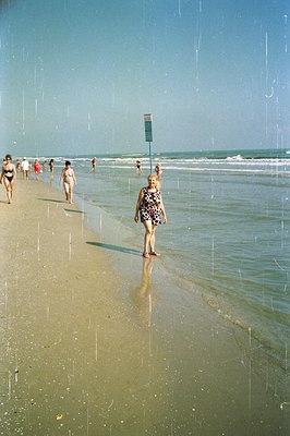 Seaside scene captures a 1960s beach vacation. A woman in a patterned bathing suit strides toward the viewer, with bathers vi...