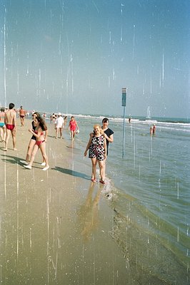 A couple strolls across a sandy beach during a sudden rainfall, surrounded by other beachgoers. The man wears sunglasses and ...
