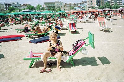 A woman seated on a green beach chair, facing the camera. Numerous beachgoers relax on colorful striped umbrellas and chairs ...