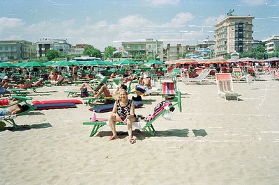 A woman sits on a sandy beach amongst rows of vibrant green and red umbrellas and deck chairs. The scene depicts a crowded Eu...