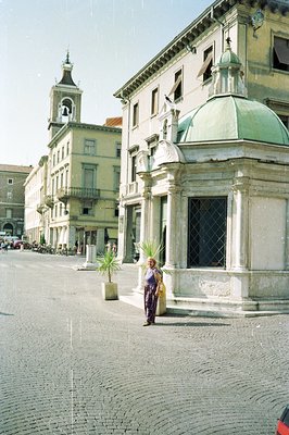 A woman stands near a classical pavilion with a green dome in a European town square. Ornate buildings with balconies line th...