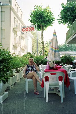 A woman seated at a table with red plastic chairs and a matching tablecloth outside Hotel GOYA, likely a seaside resort. The ...