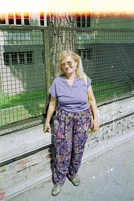 A woman stands casually in a courtyard with an older, monolithic building behind her. She wears a lavender tee, patterned wid...