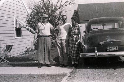 A three-person group stands beside a 1951-1953 Ford Custom sedan. A man in a work shirt and cap is flanked by a man in a t-sh...