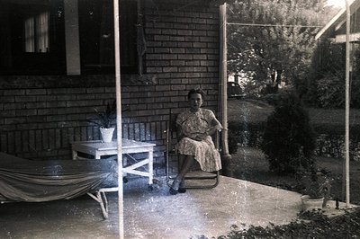 Woman seated on a porch in a vintage dress, likely 1950s. Features a brick facade, lawn, and foliage. A folding chair and sma...
