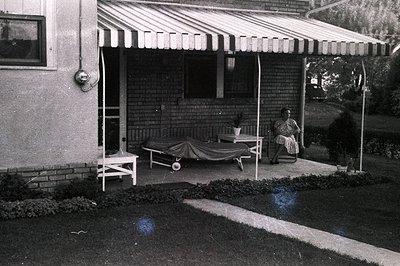 A wide, striped awning shades a brick home's porch. A woman sits on a chair near a chaise lounge, both pieces of furniture ap...