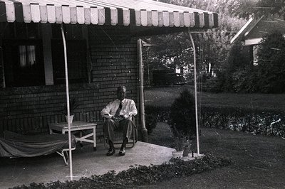 A man seated in a rattan chair on a brick porch, overlooking a shaded garden. The architecture suggests mid-century American ...