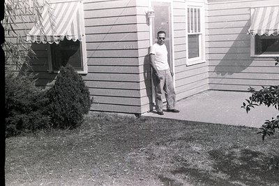 A man in a short-sleeved shirt & trousers leans against a clapboard house with awnings. The address "2831" is visible on the ...