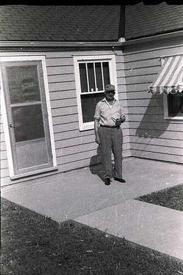 Man in a short-sleeved shirt and cap stands on a concrete porch outside a single-story home with a small awning. Likely a 195...