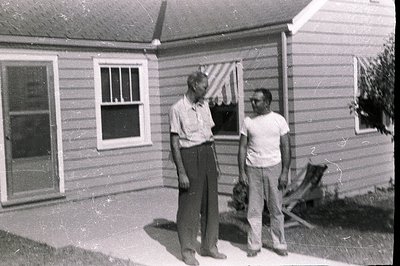 Two men stand outside a modest, single-story home with clapboard siding and a screened porch. The scene suggests a rural or s...