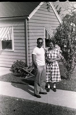 A young couple stands proudly outside a charming, low-slung home with striped awnings. The man wears sunglasses and a white t...
