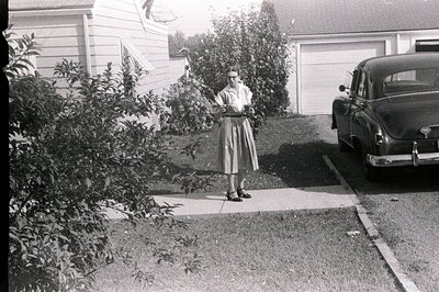 A young woman in a full skirt and blouse stands on a concrete walkway, posed next to a vintage car (likely a 1951 Oldsmobile)...