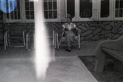 A woman sits in a wrought-iron chair outside a stone building with numerous windows. She wears a dark, knee-length dress and ...