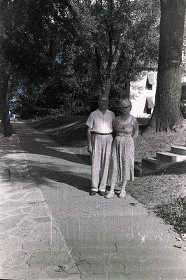 A couple, a man in trousers and a woman in a floral dress, stand on a paved walkway near a building with a prominent gable. M...