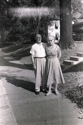 A formally posed black-and-white portrait shows an elderly couple standing on a paved walkway in front of a large, multi-stor...