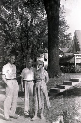 Three formally-dressed individuals stand on a walkway in front of a large tree and a two-story, gabled home. The scene sugges...