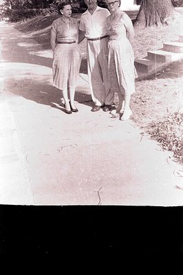 A monochrome image depicts a group of three people walking along a path. Two women and one man are dressed in mid-century att...
