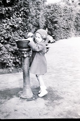 A young girl in a coat and hat leans on an ornate, aged fountain in a park setting. The fountain's basin shows signs of rust....